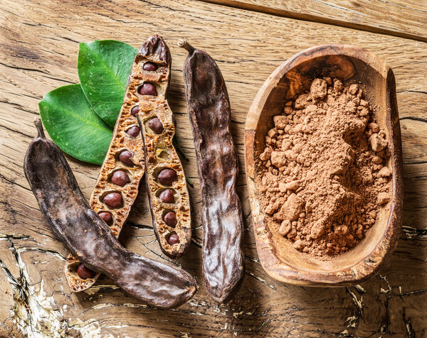 Algarroba en polvo en un cuenco sobre una mesa de madera junto a vainas de algarroba